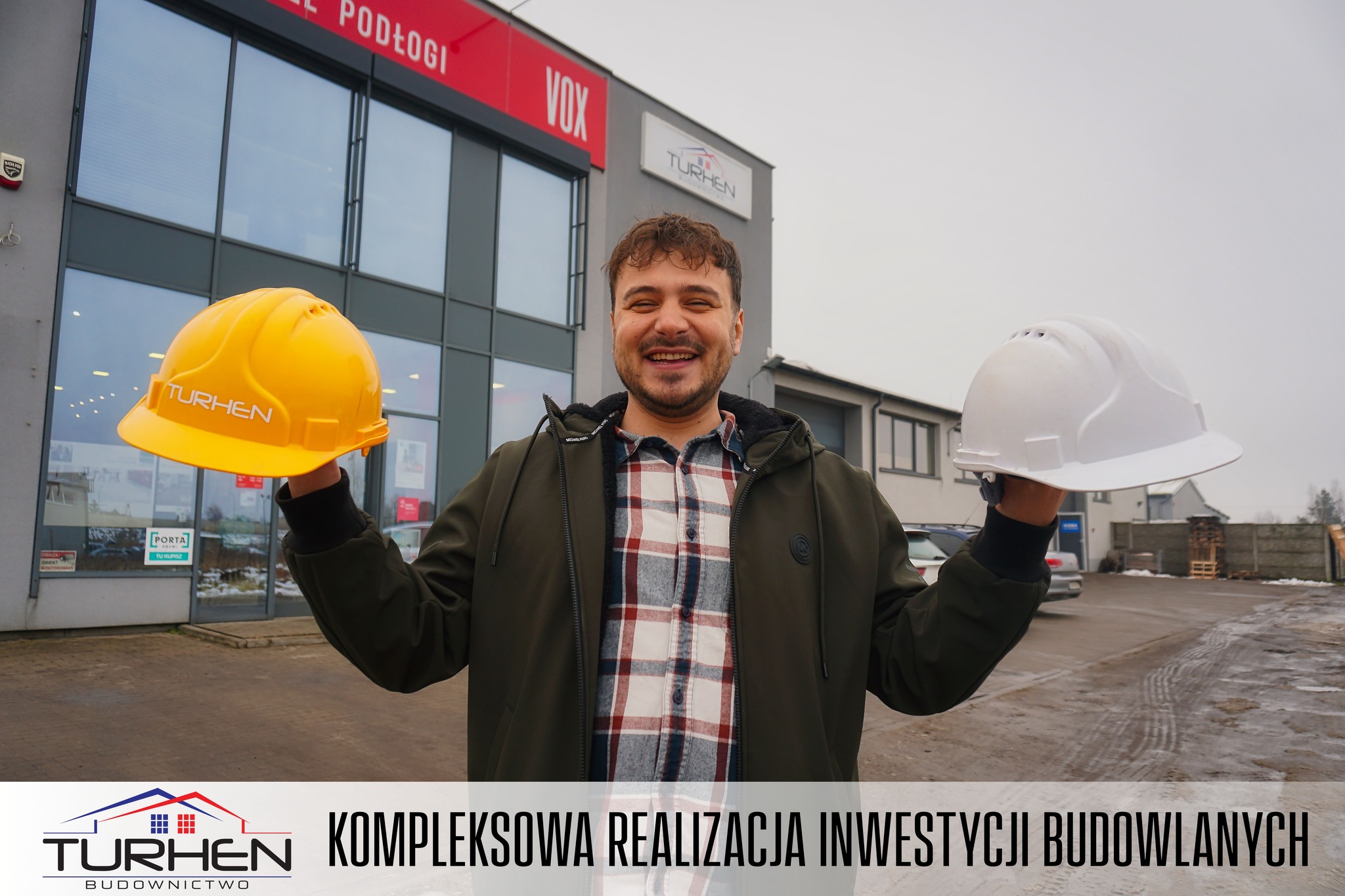 Bartek z Turhen Budownictwo trzyma dwa hełmy budowlane przed siedzibą Turhen Budownictwo, symbolicznie łącząc pracę w biurze i na budowie.